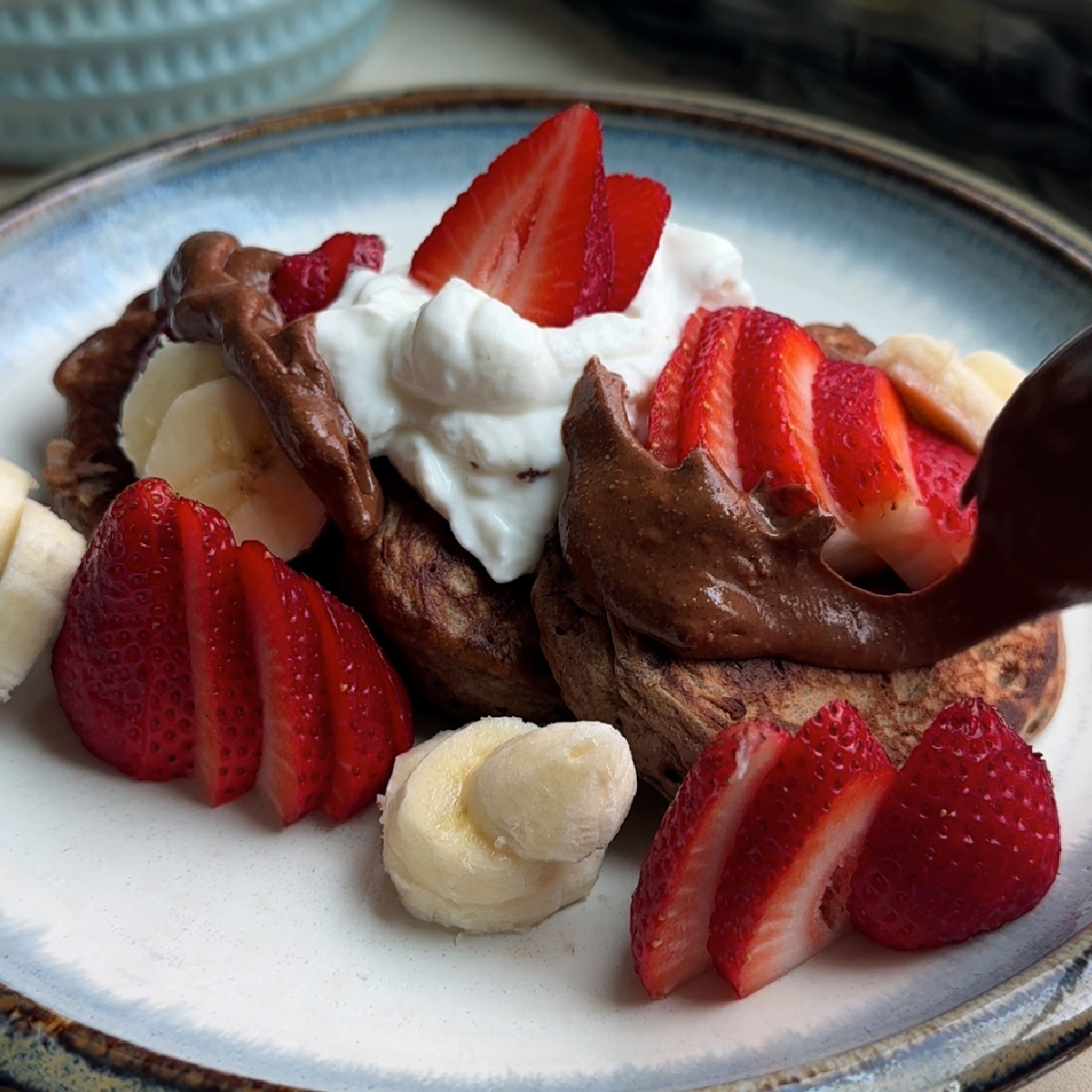 three chocolate pancakes with greek yogurt dolloped on top, topped with sliced strawberries and bananas and Nutella